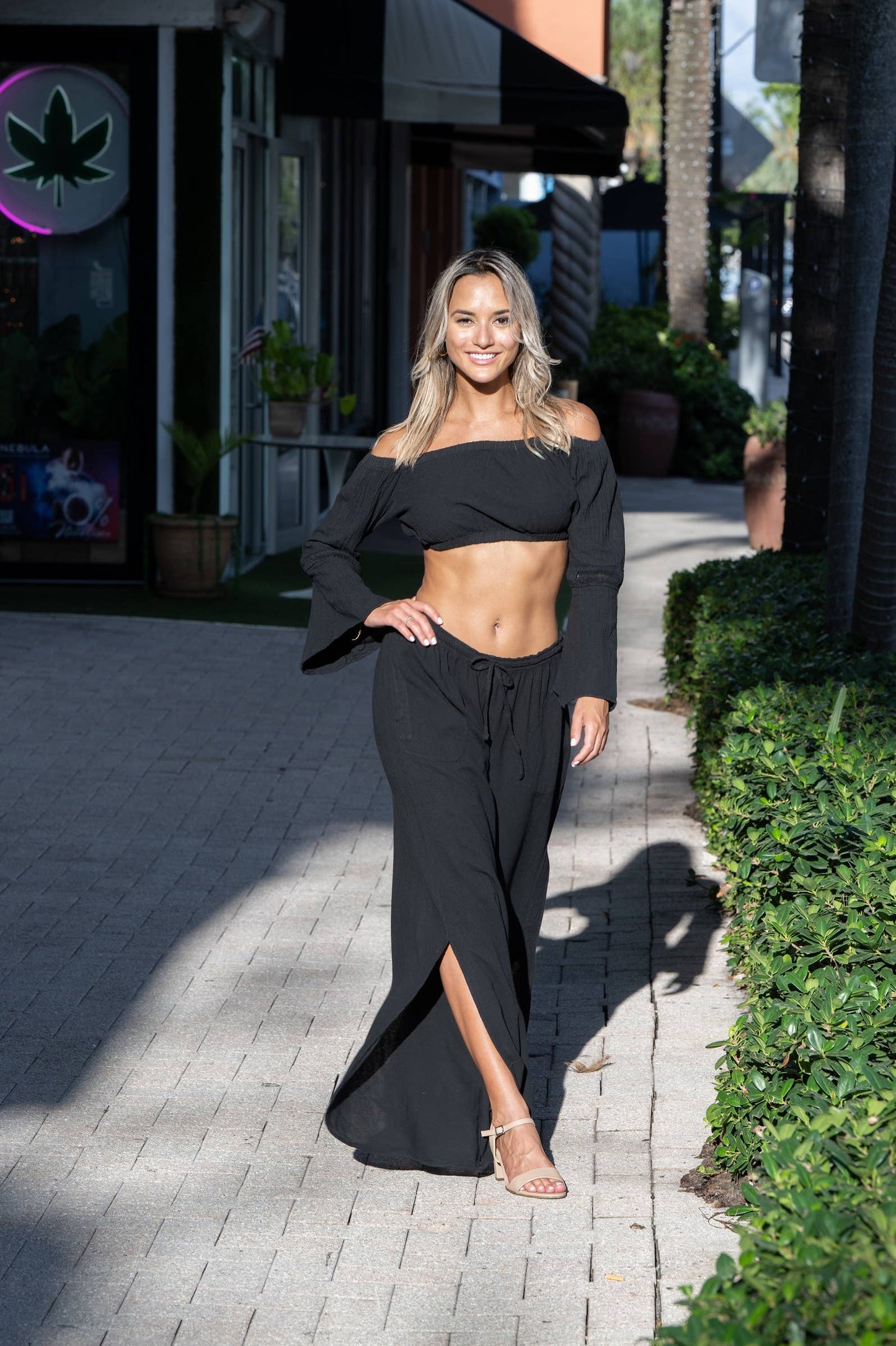 Woman in a black outfit standing on a sidewalk with greenery and a building in the background