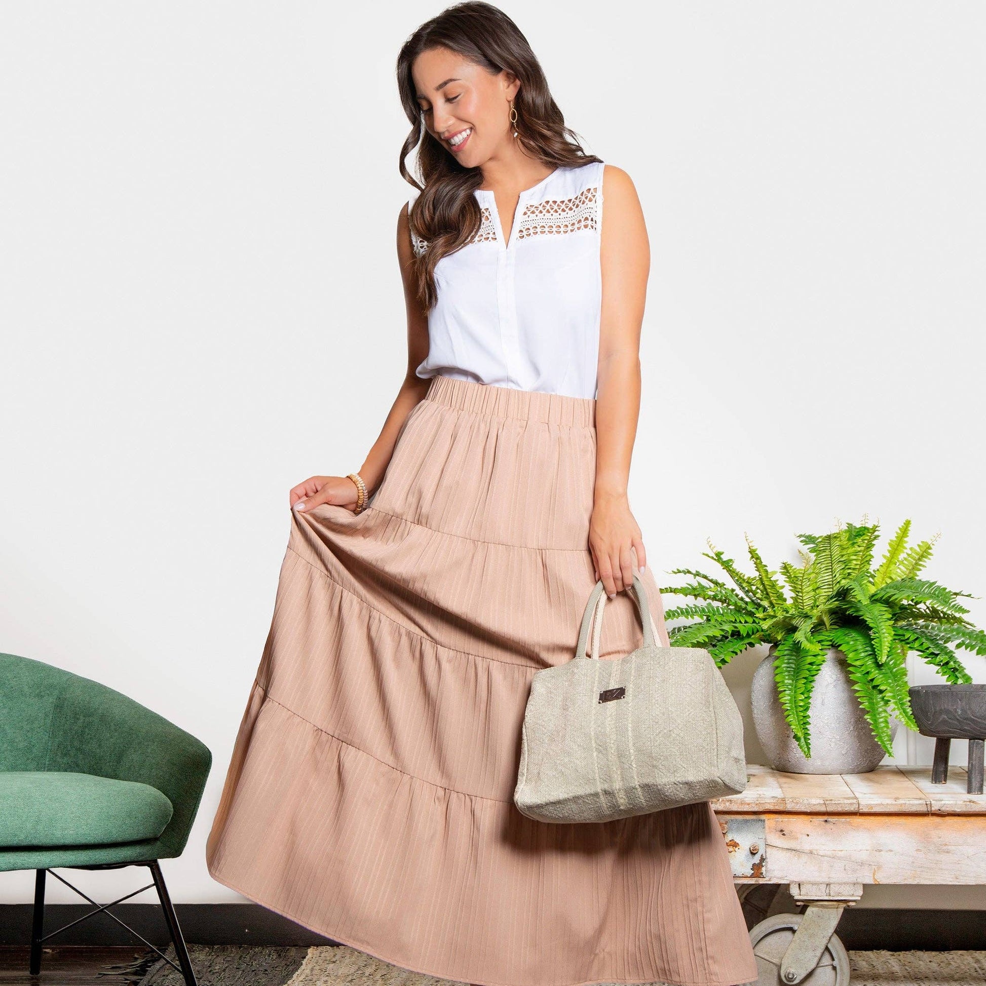 Woman in a white top and beige skirt holding a handbag in a room with a green chair and potted plant.